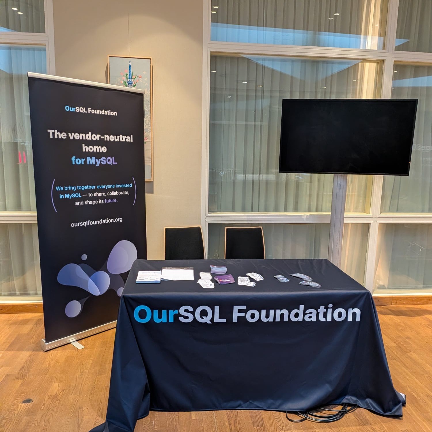 The OurSQL Foundation booth at Percona Live Amsterdam, with a roll-up banner reading 'The vendor-neutral home for MySQL' next to a table of flyers and stickers.